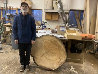A man stands smiling next to a cross-section from a spruce tree. Standing on edge, it comes up to his waist.