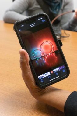 Hand holds up phone with image of two hoop lights. The light on the left is blue-green and smaller, whereas the right side light glows red and is larger. The lights sit against a wall, next to each other, and reflect their colors off the floor,