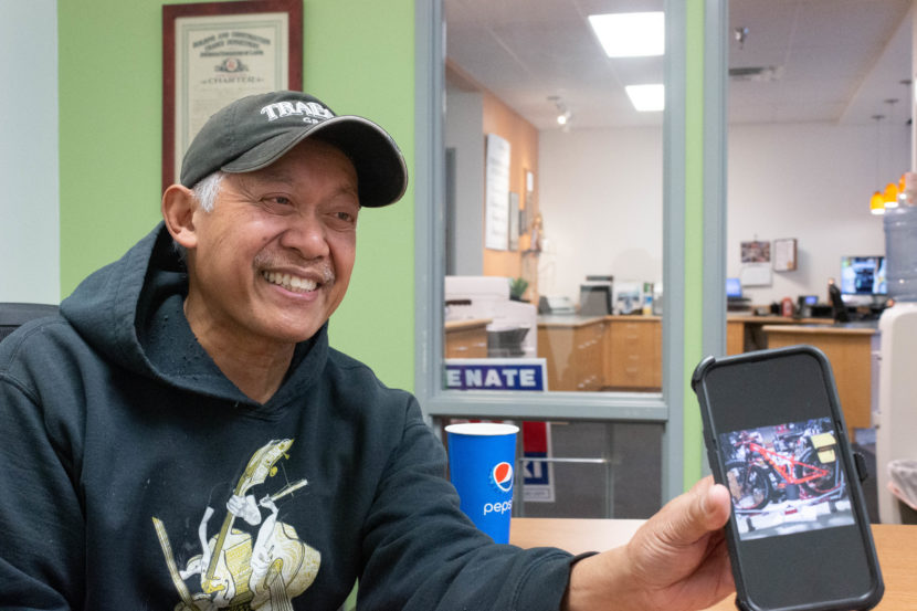 Man smiles while holding a phone up inside a green room. The phone shows an image of a red motorbike after repair.