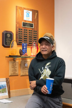 Man speaks while gesturing with his hand and holding a Pepsi cup. He is near a shelf with many awards and plaques, including two of his own.