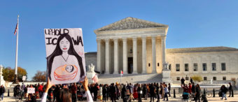 A group gathered in front of the U.S. Supreme Court building. A pair of hands is holding up a sign that says "Uphold ICWA"
