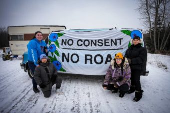 Four people on a snowy road holding a sign that says no consent, no road