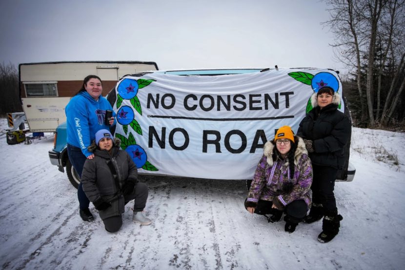 Four people on a snowy road holding a sign that says no consent, no road
