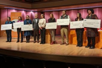 Nine people stand on a stage, seven of them holding giant checks.