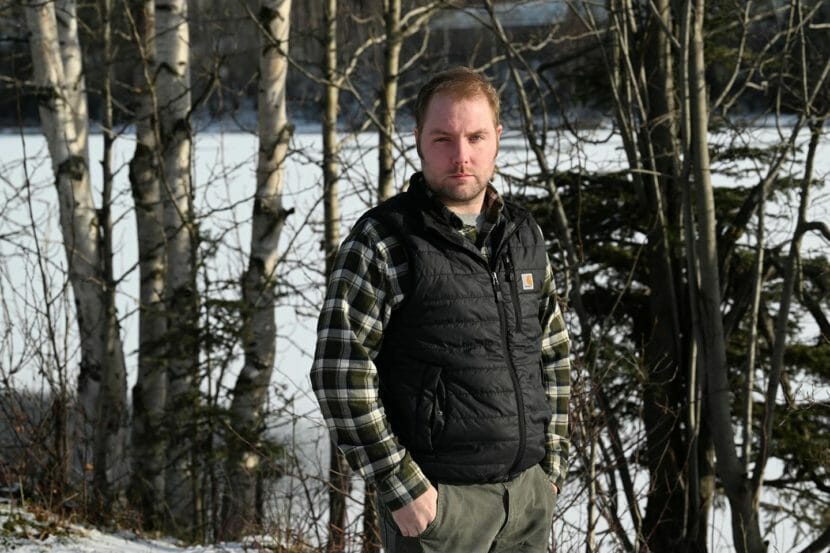 A man in a Carhartt vest and a plaid shirt stands in the woods.