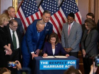 Lawmakers stand around Nancy Pelosi, who is seated and signing a bill on a blue desk with a "The respect for marriage act" sign on it.