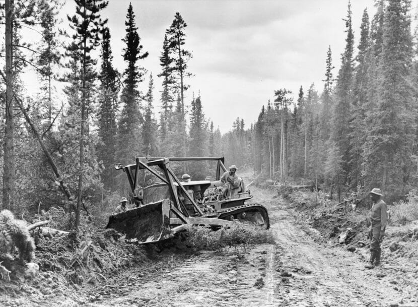 A black and white photo of two men using a bulldozer to build road through boreal forest.