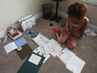 A young woman sits on the floor surrounded by paperwork.