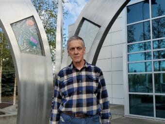 A man in a plaid shirt stands outside a glass-and metal building facade.