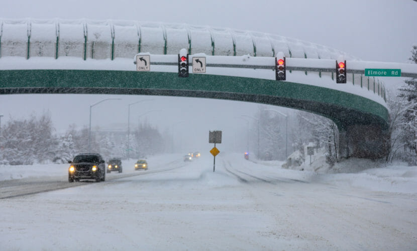 Cars driving on snow-covered roads, with heavy snow still falling.