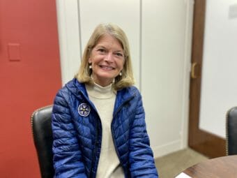 Lisa Murkowski, smiling, sits in a chair wearing a blue, puffy jacket