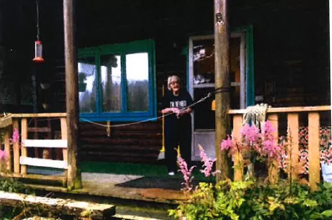 A woman stands on a rustic porch with fireweed blooming in front of it.