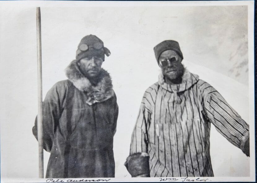 A black-and-white photo of two men in historic winter gear with snow goggles standing on a mountain.