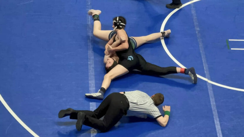 A ref lies on a wrestling mat and watches as one wrestler pins another