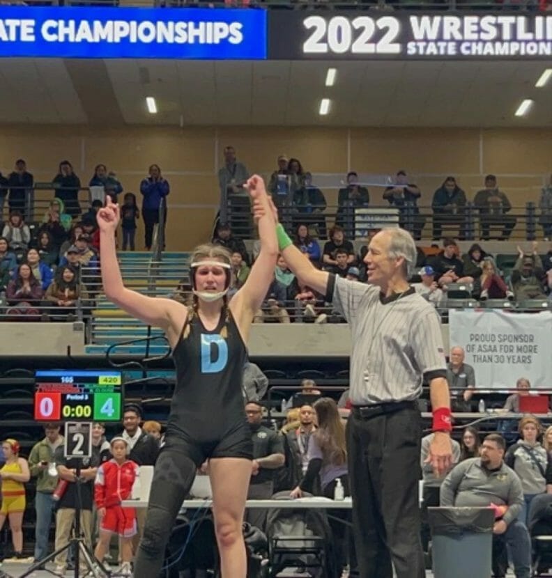 A smiling referee holds a girl's fist aloft as she stands on a wrestling mat