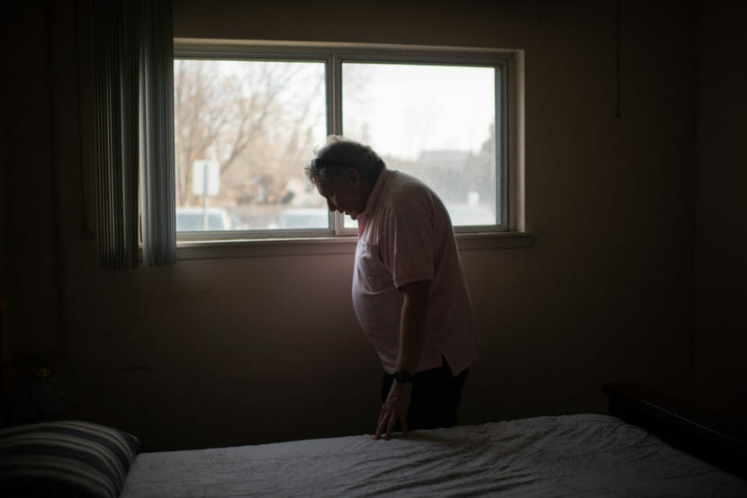 An older man stands by a window in a bedroom with light streaming in.