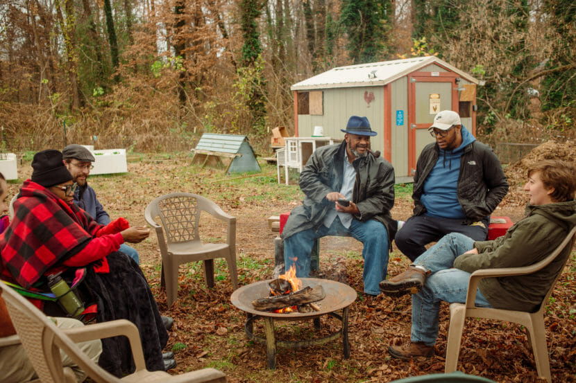 Five people sit talking around a fire pit on picnic chairs.