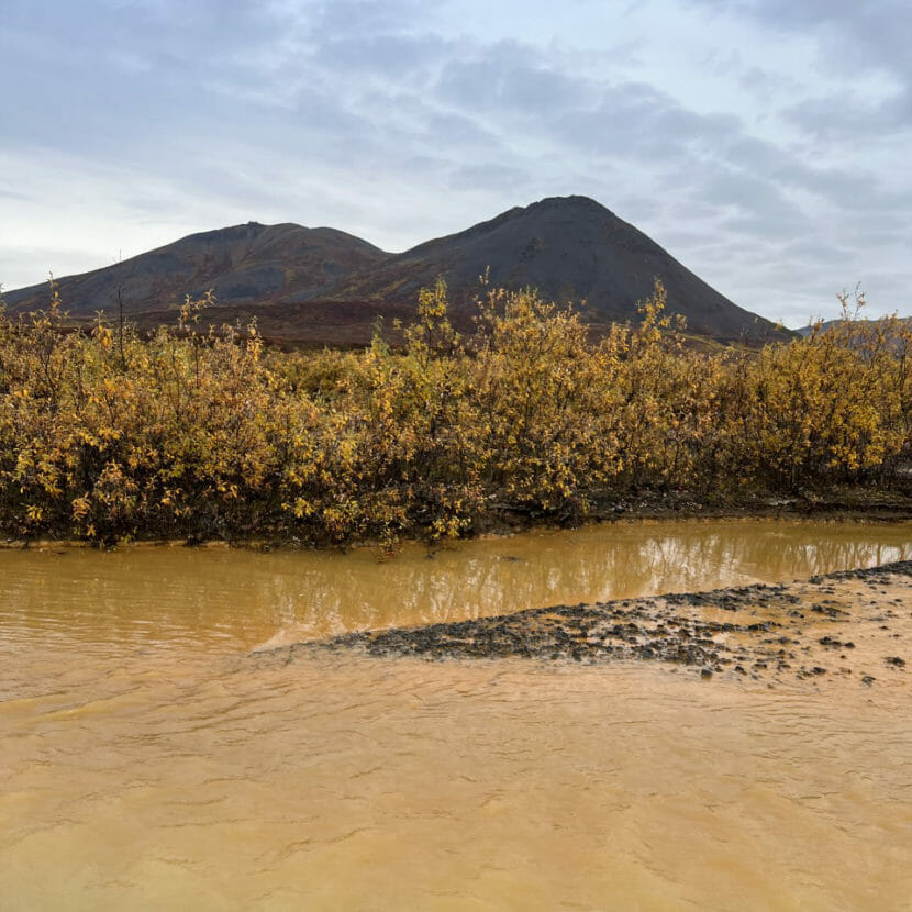 A river running orange with orange brush on its bank and bare hills in the background.