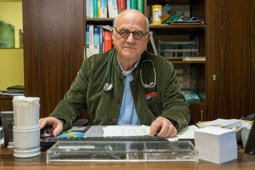 A man sits behind a desk wearing a stethoscope around his neck.