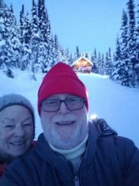 A selfie taken by an older couple in winter clothes with a log cabin in the background