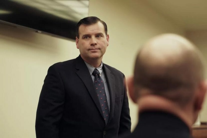 A man in a coat and tie stands in a courtroom.