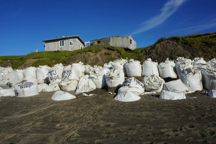 A wall of large sand bags along a beach, with houses in the background.