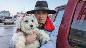 A man in a wide-brimmed black hat holds a white fluffy dog while leaning against a red pickup.