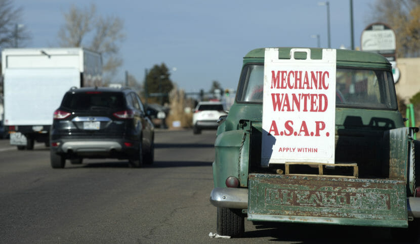 A sign advertising the need for a mechanic stands in the back of aged Chevrolet pickup truck.