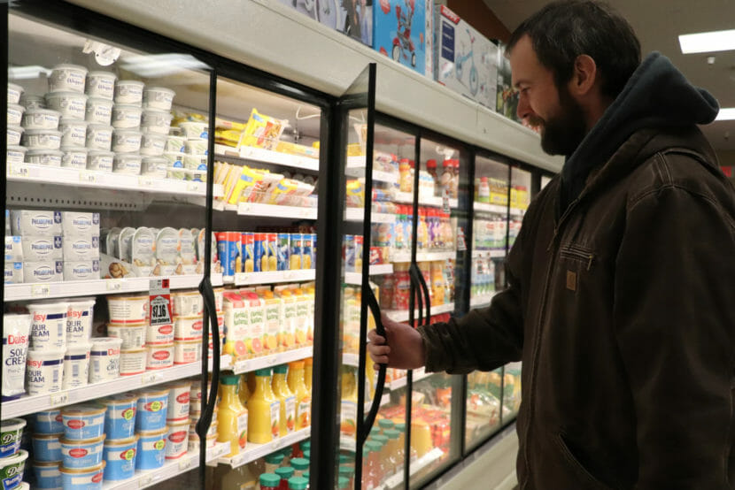 A bearded man holds a door open in the refrigerator section of a grocery store