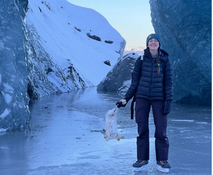 A young woman in winter clothes stands on ice dangling a dead hare from her hand.