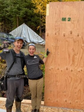 A man and a woman stand side-by-side in construction gear. They are smiling and standing near an upright wood panel.