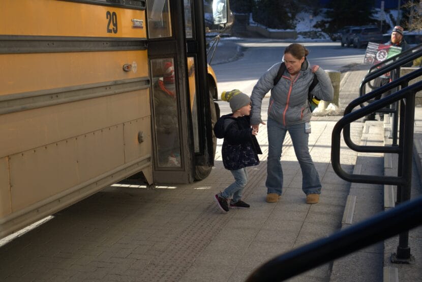 A child in a black jacket and gray hat holds an adult's hand while exiting a yellow school bus.