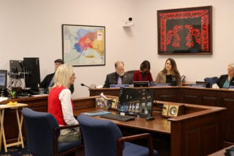 Deena Bishop wears a red sweater vest and sits on a blue chair in front of legislators.