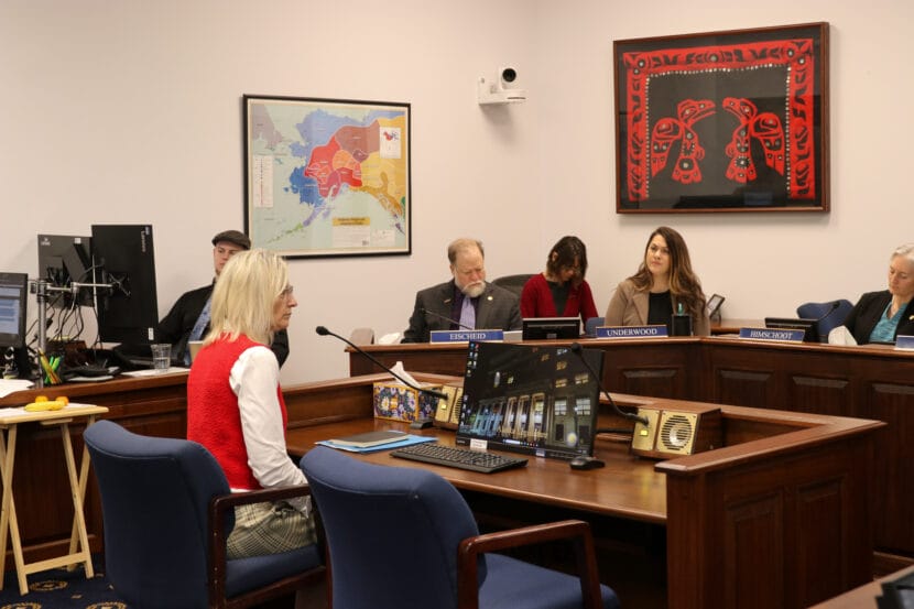 Deena Bishop wears a red sweater vest and sits on a blue chair in front of legislators.