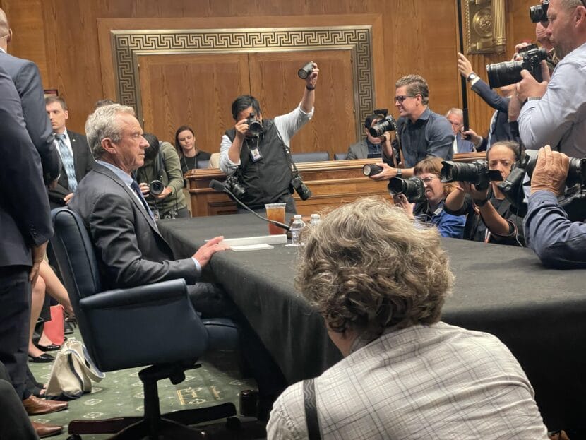 Besuited man at a table, surrounded by photographers.