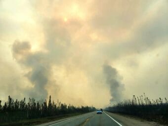 A line of cars travels on a two-lane highway between burned spruce forest with columns of smoke rising on either side.