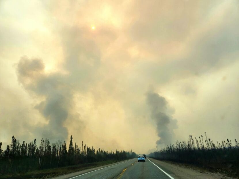 A line of cars travels on a two-lane highway between burned spruce forest with columns of smoke rising on either side.