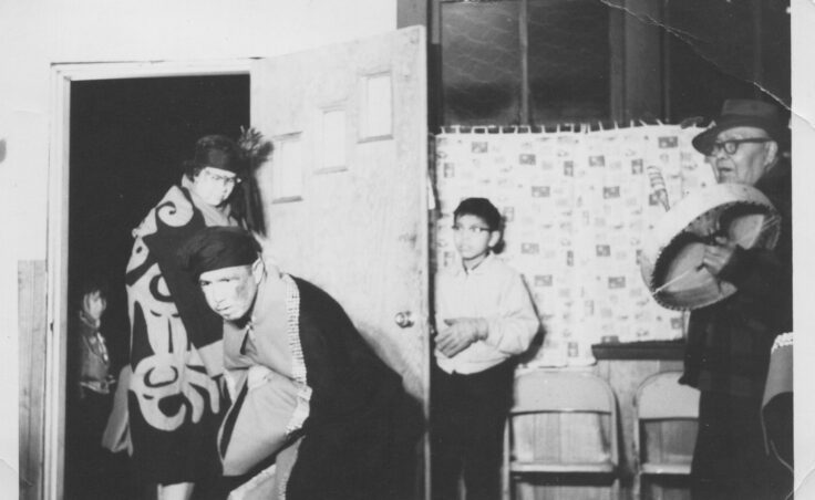 Dancers in regalia dance into a room, as a man drums and a boy looks on. From the Cyril George Photo Collection.