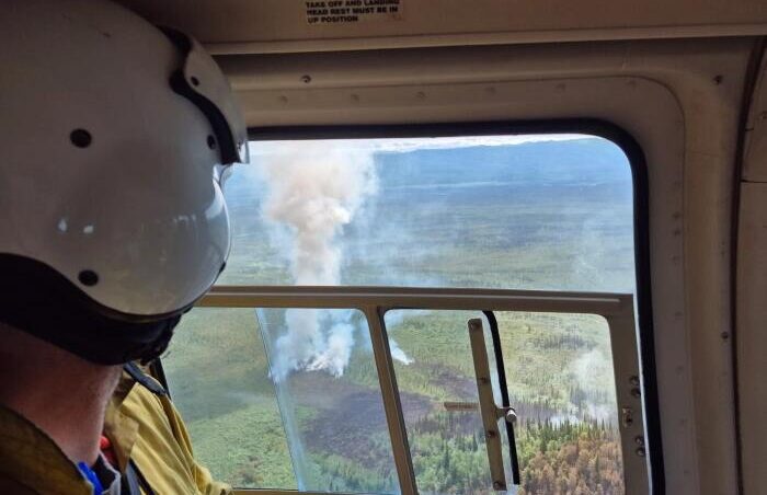 A man in a flight helmet looking through an aircraft window at a wildfire below.