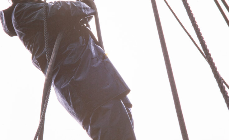 A boy in a red bandana and Xtratufs smiles down at the camera as he swings from a rope on a pulley system. From the Cyril George Photo Collection.