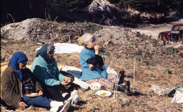 Three old women sit on a beach, having a picnic. From the Cyril George Photo Collection.