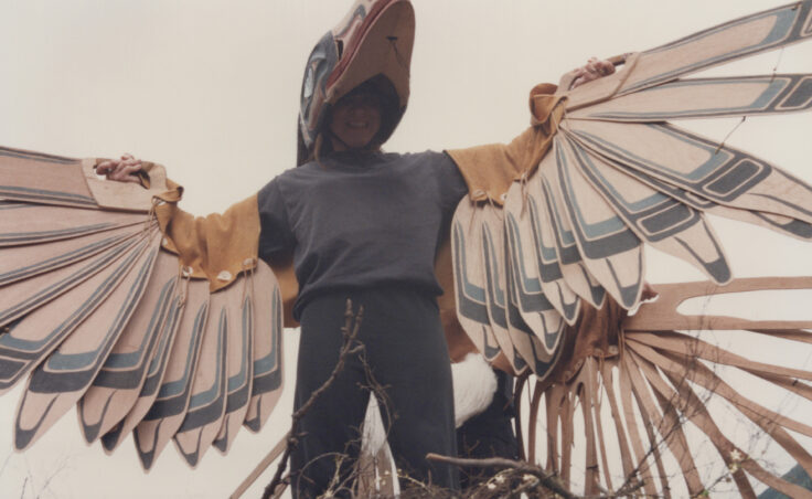 A person smiles from under what looks like a formline Raven headpiece and wings. From the Cyril George Photo Collection.