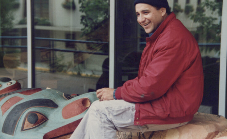 A smiling man sits on a partly carved and painted kootéeyaa. From the Cyril George Photo Collection.