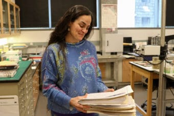 A woman with long brown hair and a blue sweater smiles as she looks at a stack of manila folders.