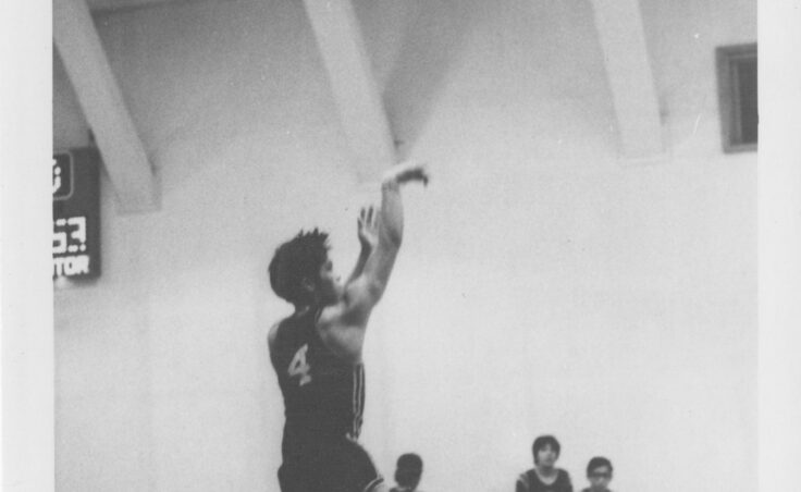 A boy shoots a basketball as his teammates look on. From the Cyril George Photo Collection.