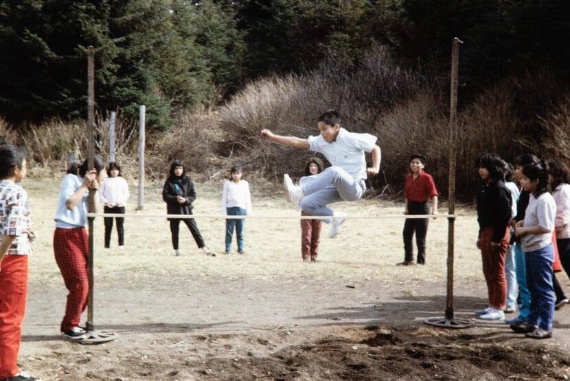 A boy jumps over a bar as his peers look on. From the Cyril George Photo Collection.