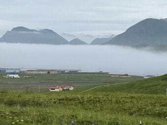 buildings sit on a green slope overlooking a large bay with mountains behind it