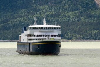 A blue-and-white Alaska ferry traveling through an inlet, with steep forested hills in the background.