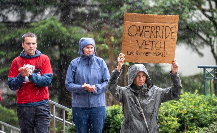 Suzanne Cohen holds a sign in the rain outside the Alaska Stae Capitol on Friday, Aug. 1 calling on lawmakers to override Gov. Mike Dunleavy's veto of more than $50 million in public school funding.