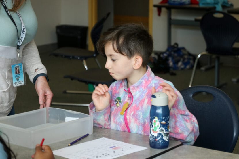 A child in a fleece jacket looks at a translucent plastic container with her hands hovering over a small water bottle. 